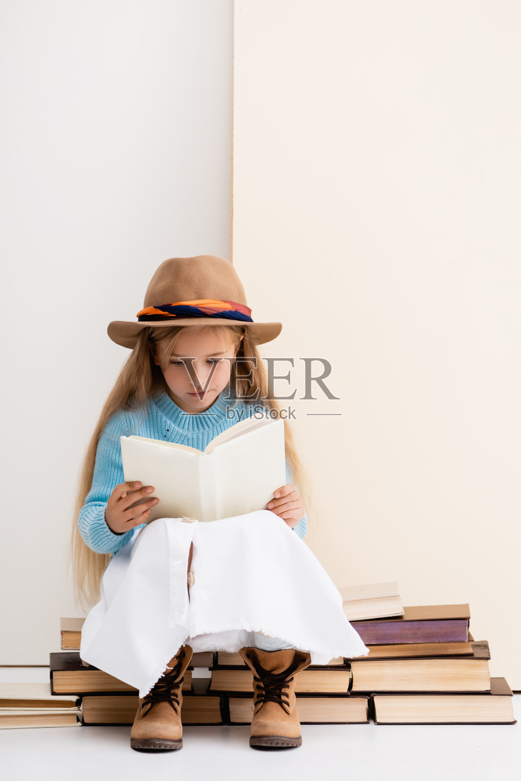 时髦的金发女孩戴着棕色的帽子和靴子,白色的裙子和蓝色的毛衣坐在古董书和阅读附近的米色墙壁照片摄影图片