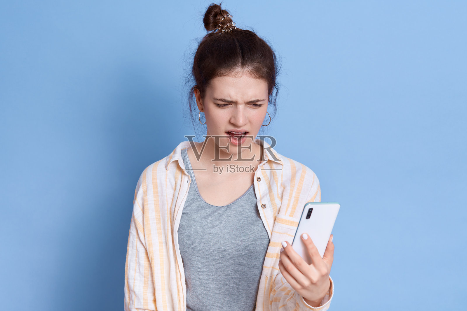 迷人的年轻褐发女人感到怀疑和困惑,不知道该做什么决定,使用智能手机,保持嘴巴张开,摆出孤立的蓝色背景。照片摄影图片