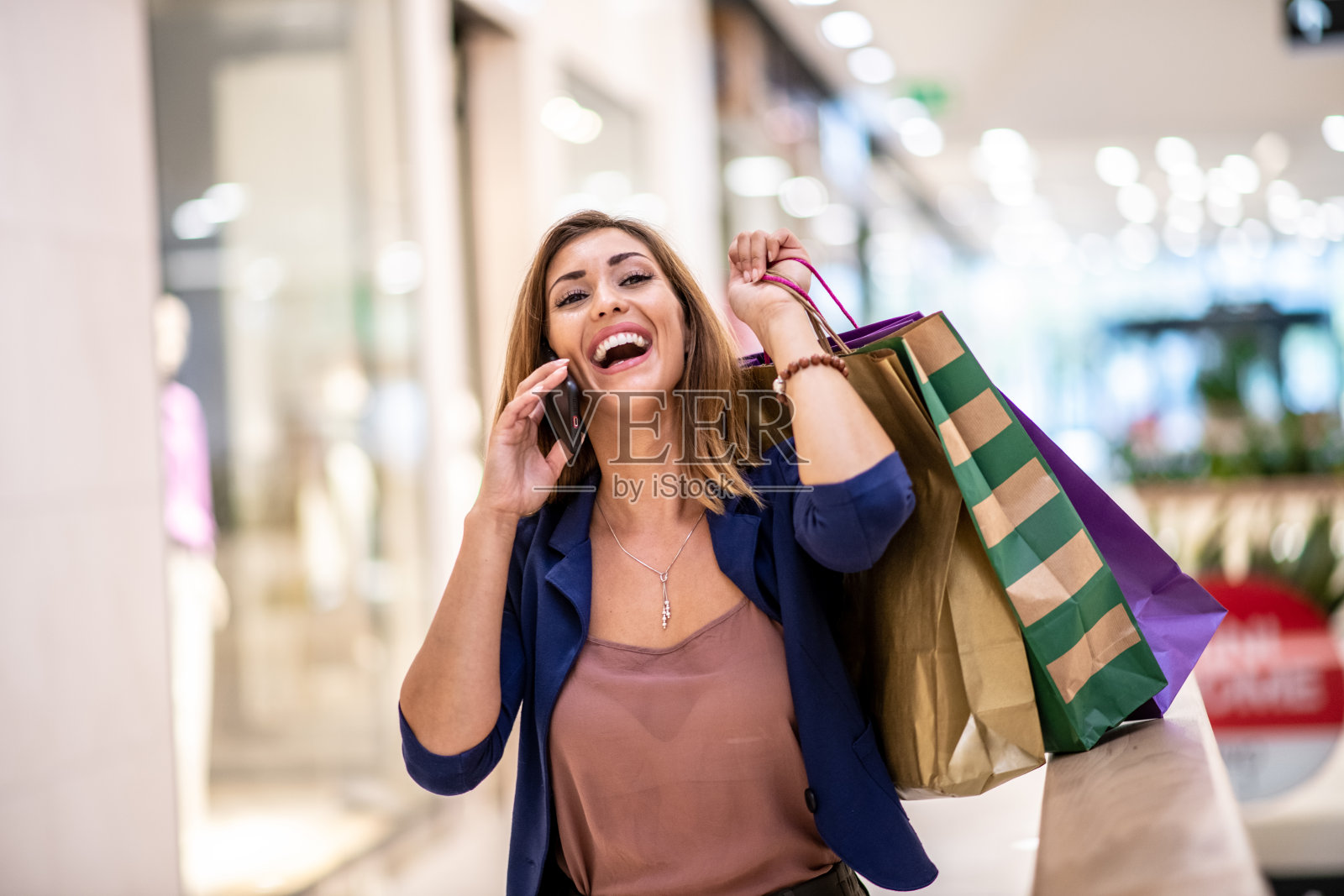 年轻漂亮的微笑开朗的时尚女人站在购物中心,拿着包和讲电话。她正在和她的朋友谈话。照片摄影图片