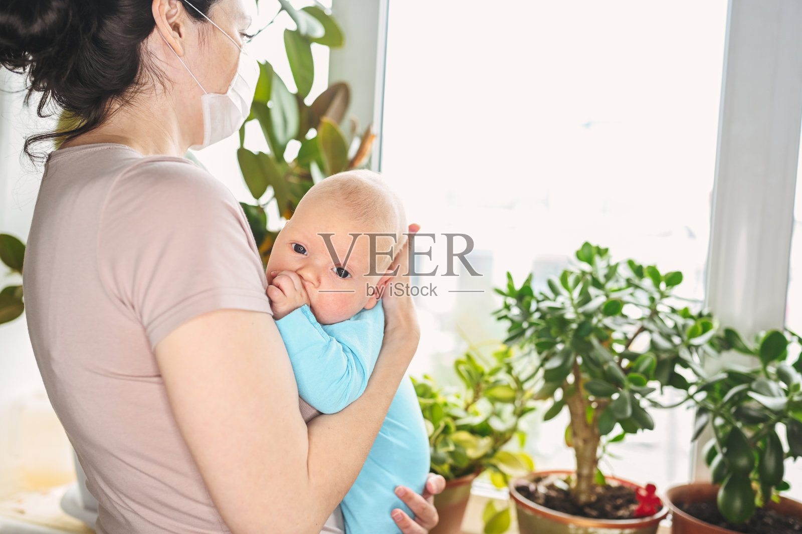 美丽的母亲戴着防护口罩,抱着她可爱的新生儿子,保护他免受病毒和感染。冠状病毒covid-19预防。居家隔离。家人在家靠窗。照片摄影图片