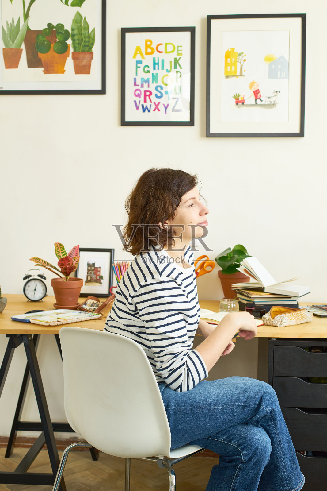 女性艺术家在她的工作场所在家工作。穿着牛仔裤和条纹衬衫的女人坐在桌子前画插图。寻找灵感概念。照片摄影图片