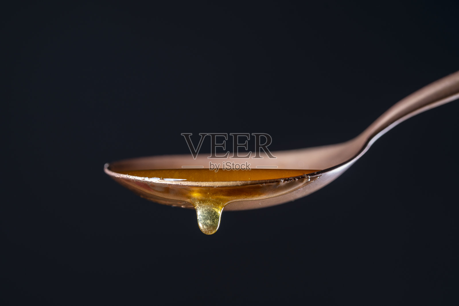 浓蜂蜜从一个金属勺子滴下,黑色背景,特写。蜂蜜从匙中流出照片摄影图片