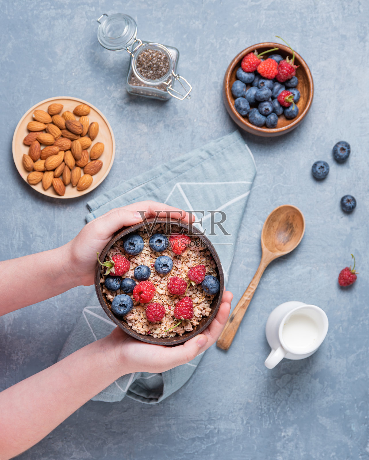 手孩子保持椰子碗与麦片,杏仁坚果和浆果在蓝色背景照片摄影图片