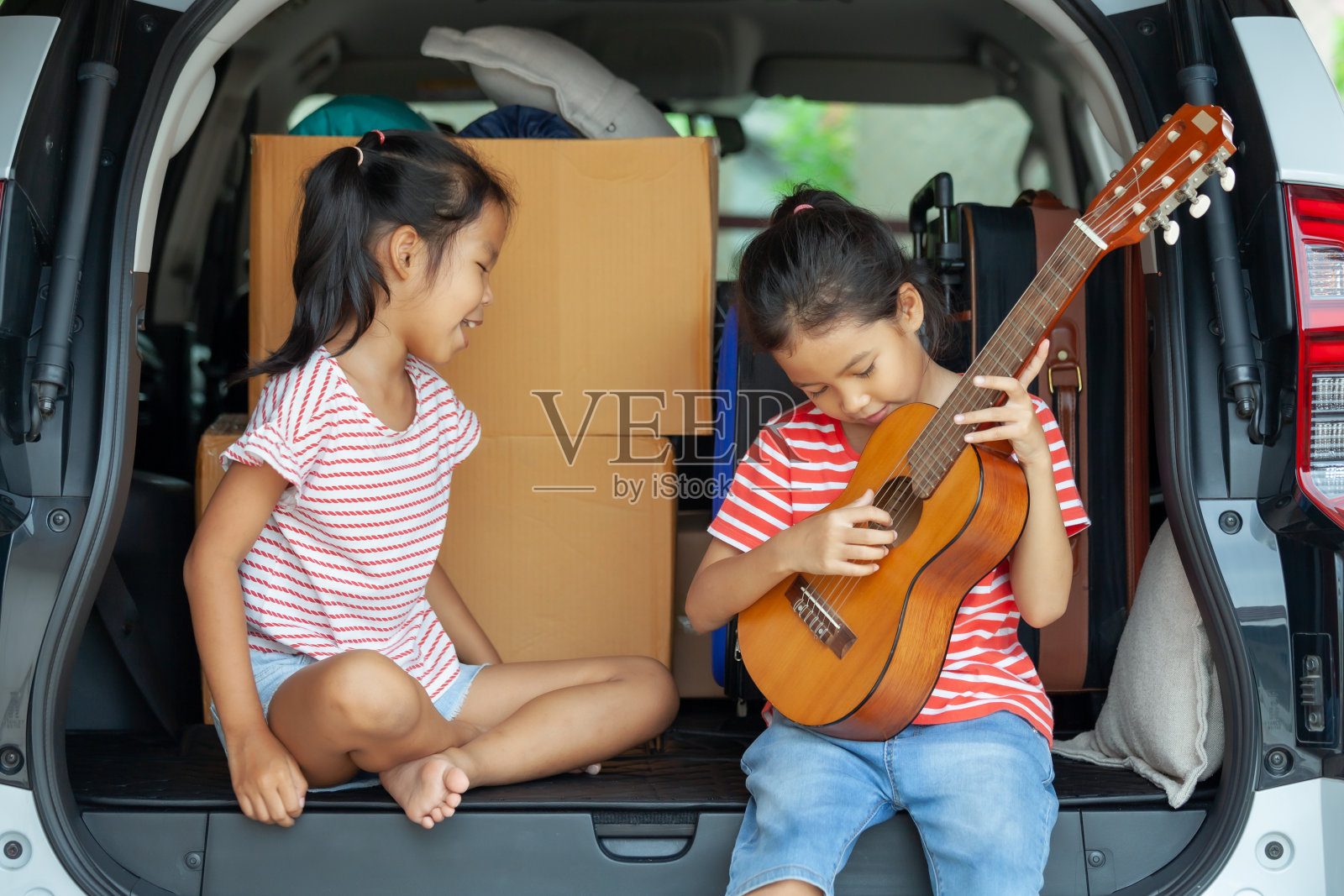 快乐的亚洲女孩弹吉他和唱歌与她的妹妹在汽车后备箱在家庭旅行。照片摄影图片