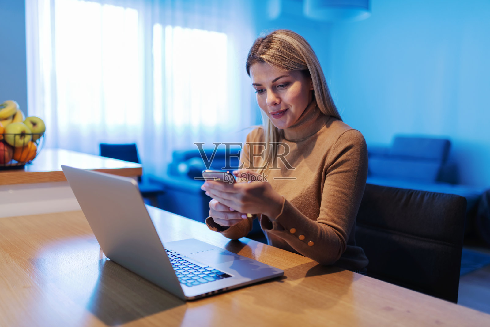 年轻漂亮的金发女商人坐在家里,在冠状病毒爆发期间使用智能手机。桌子上有一台笔记本电脑。照片摄影图片