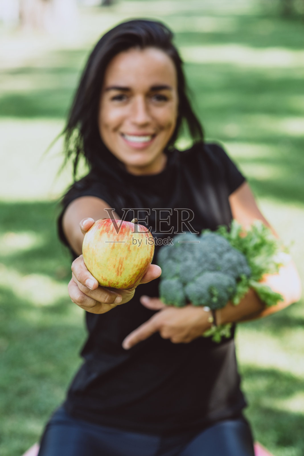微笑的年轻女子在公园里拿着一个苹果和一束绿色的花椰菜,饮食,健康的饮食。软选择聚焦。照片摄影图片
