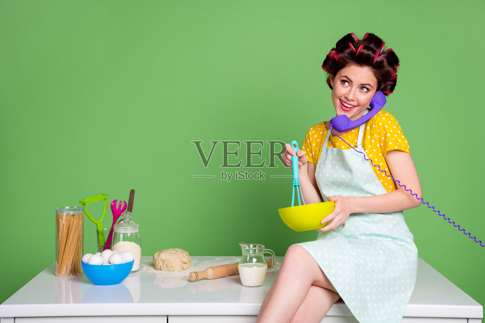 迷人的年轻女士做饭的照片,愉快的坐在桌子,厨房卷发器,头发,迷人的主妇,说话,电缆,电话,穿海报,点缀的衣服,围裙,孤立的绿色背景照片摄影图片