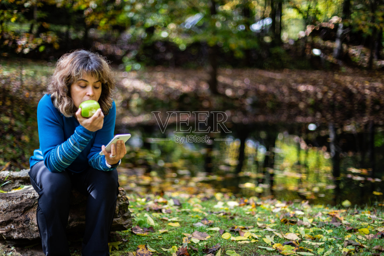 一名女子穿着蓝色毛衣,一边看手机,一边坐在石头上咬苹果,旁边的池塘被五颜六色的秋叶覆盖着照片摄影图片