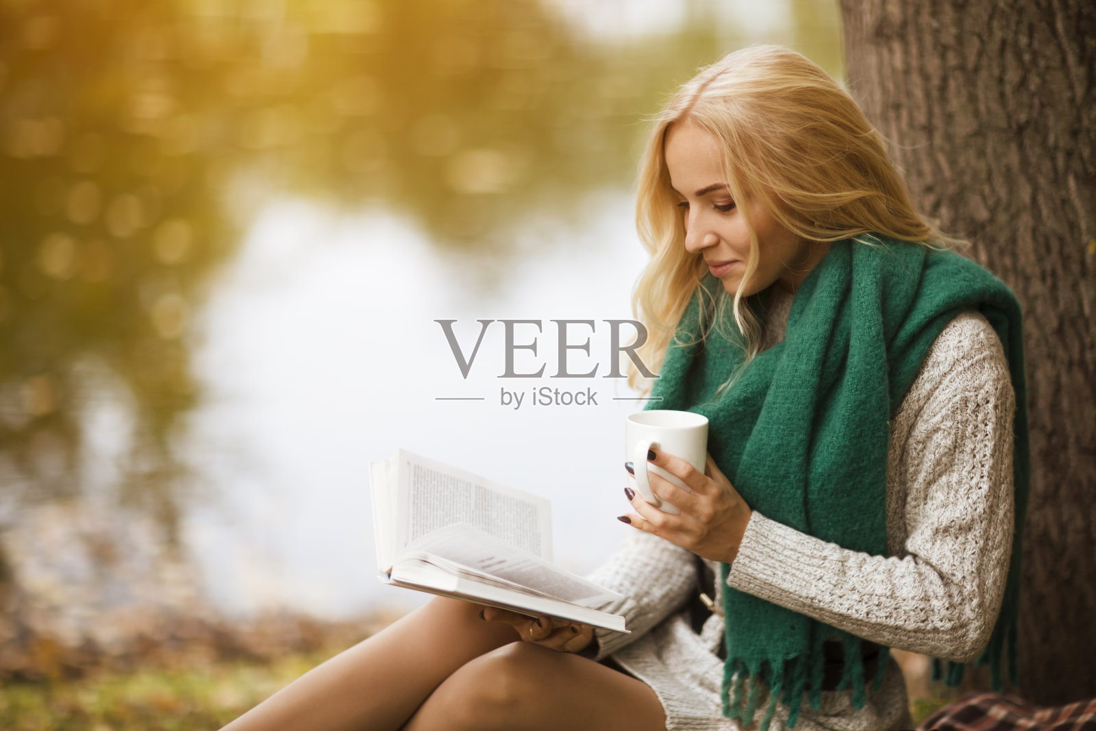 美丽的金发女郎坐在公园里的落叶上,一边看书一边喝茶照片摄影图片