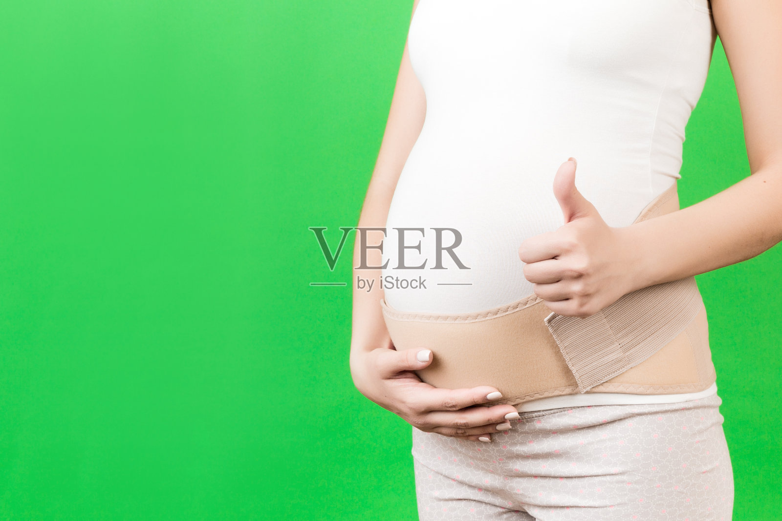 特写孕妇显示拇指向上和穿着弹性怀孕绷带,使疼痛消失在绿色背景与复制空间。骨科腹部支撑带概念照片摄影图片