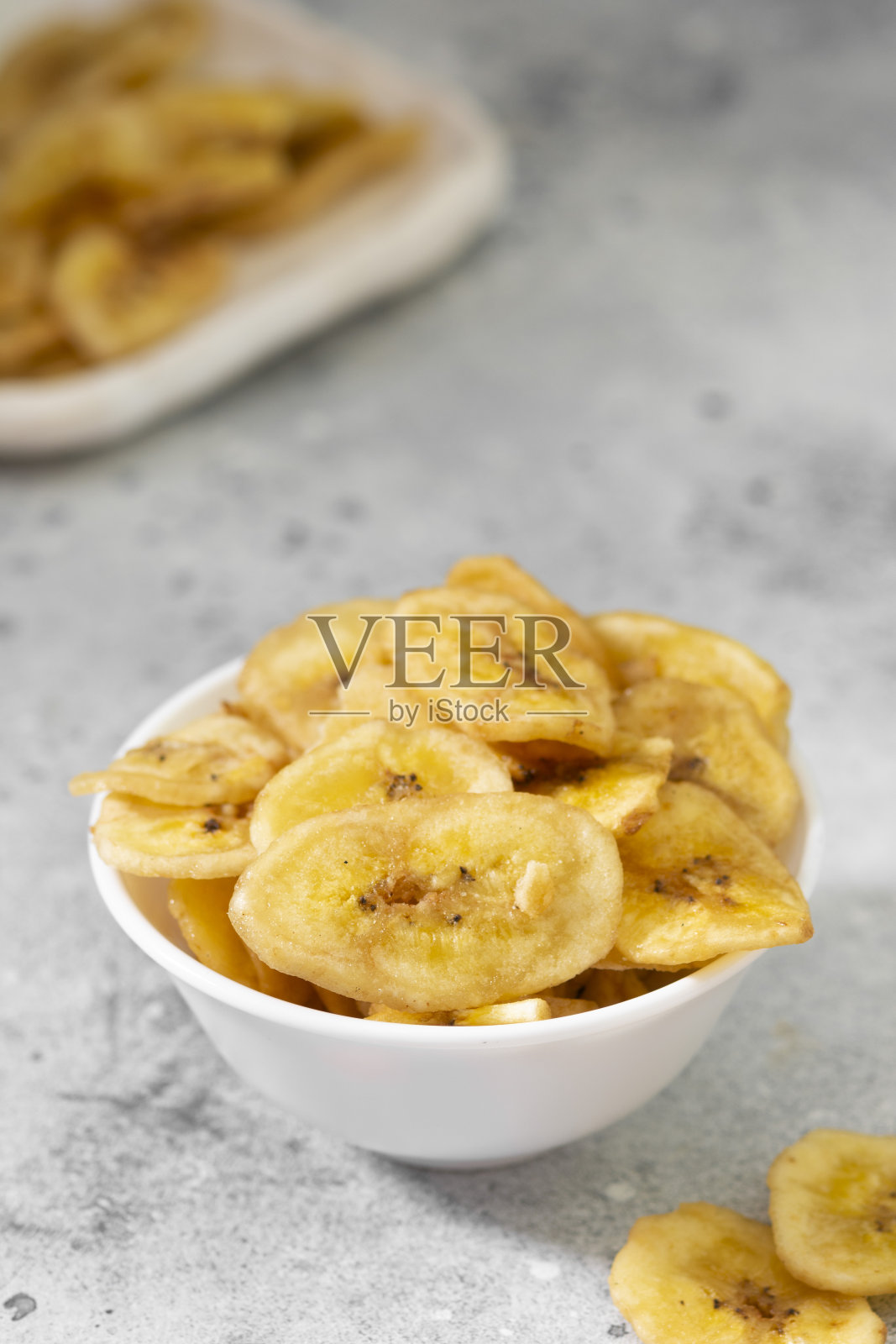 灰色的餐桌上,白色陶瓷碗里盛着干香蕉。香蕉片。适当的素食零食照片摄影图片