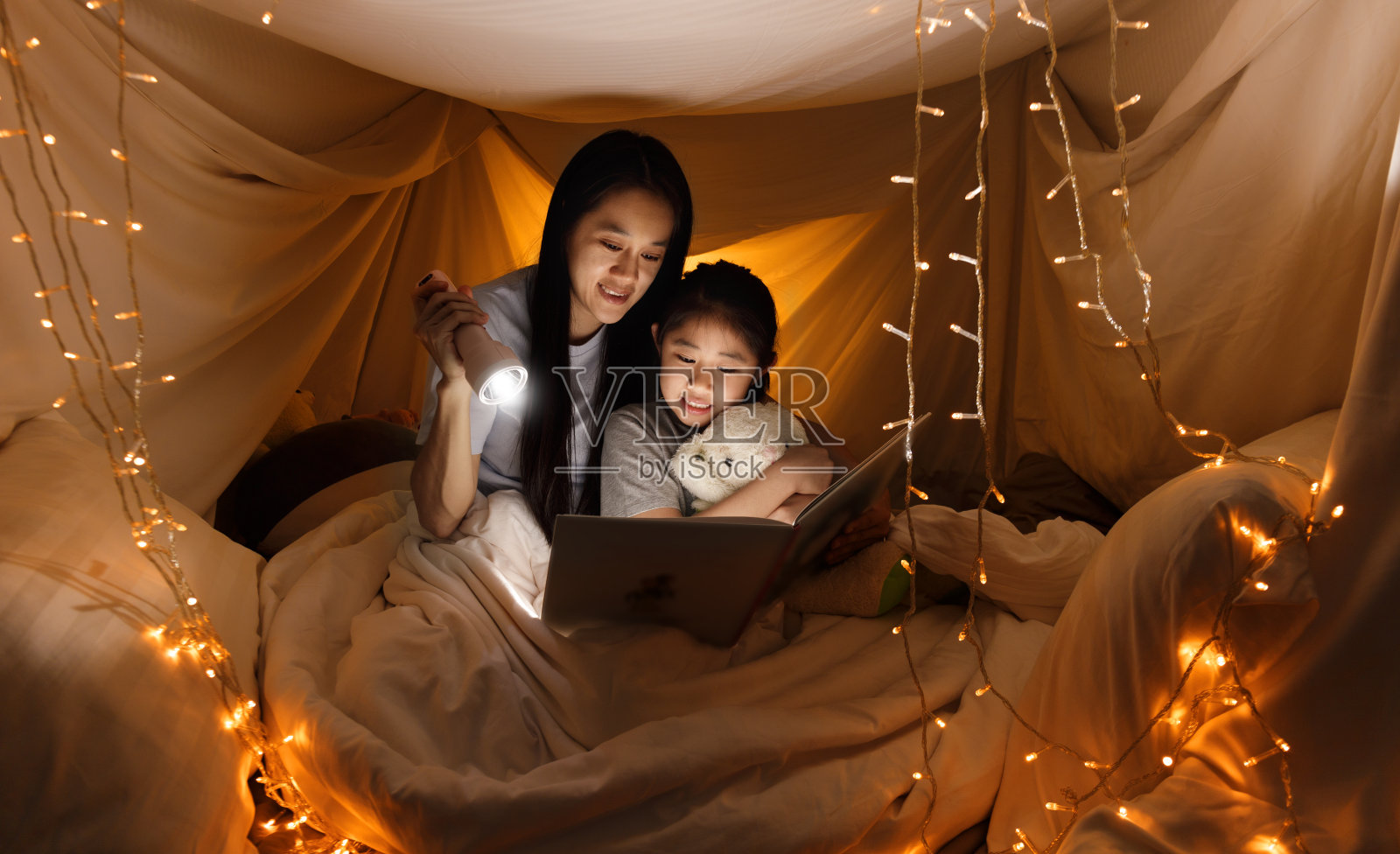 家庭的概念。姐姐和姐姐在睡觉前一起用手电看书。妹妹在床单帐篷里一起读故事书。重点选择小妹妹。带有薄膜颗粒效应照片摄影图片