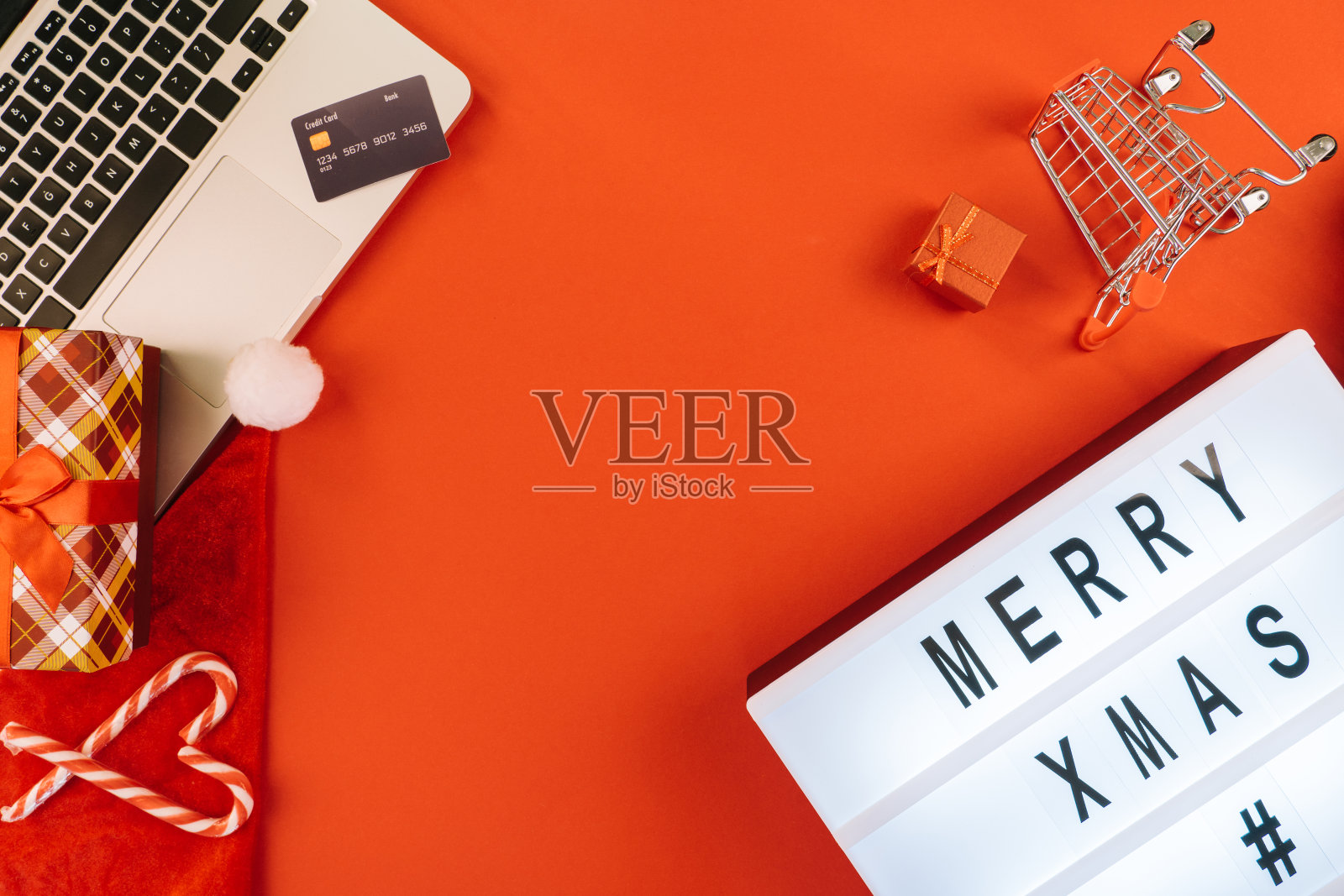 圣诞节用信用卡在笔记本电脑上购物。新年装饰品糖果手杖、购物车和礼品盒俯视图平铺在红色背景上。圣诞快乐,寒假销售理念。照片摄影图片