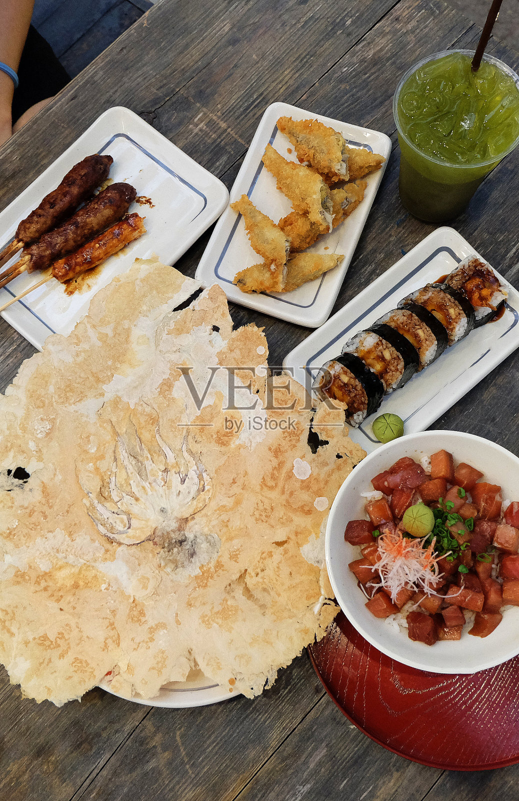 红烧鸡串、炸飞鱼、寿司卷、生鱼片、鱿鱼米饼和冰绿茶照片摄影图片