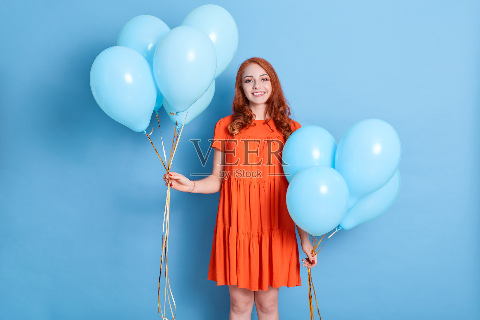 漂亮的女人气球,穿着优雅的衣服,有红色的波浪发,看着镜头,快乐的女人庆祝她的生日,孤立地站在蓝色的背景。照片摄影图片