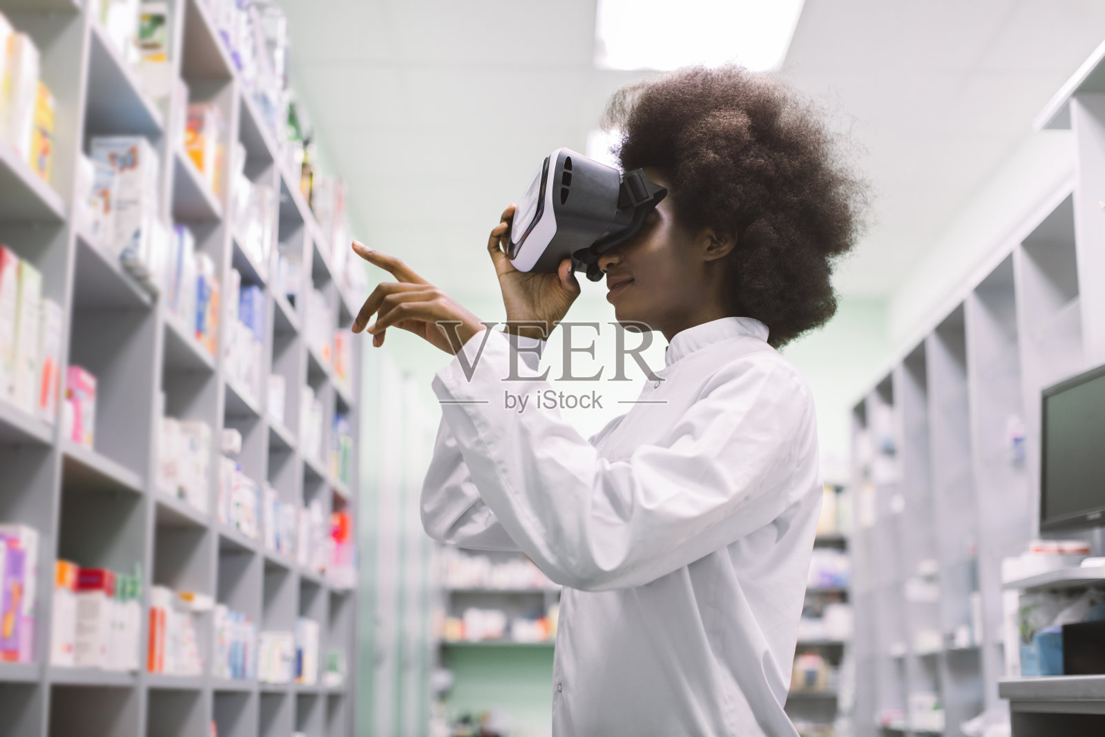 侧面的年轻职业非洲女性药剂师,戴着虚拟现实耳机,在现代药店工作,看着货架上的药品和手势照片摄影图片