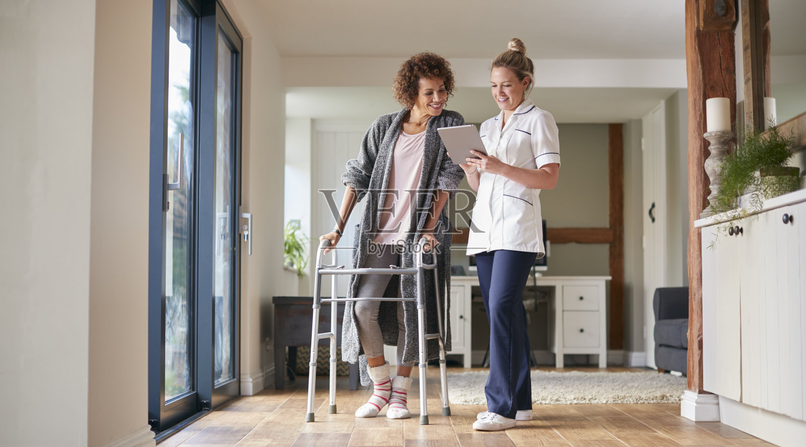 成熟的女性穿着睡袍使用步行架由女性护士帮助与数字平板电脑照片摄影图片