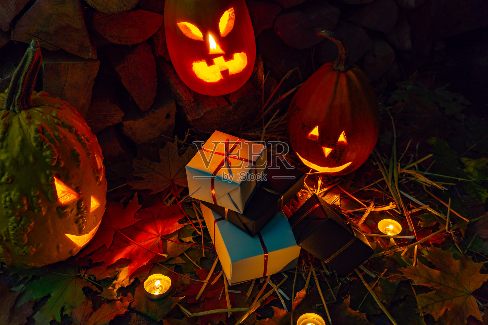 三个发光的南瓜和一串黑色和白色的礼物在秋天的树叶和蜡烛在万圣节之夜。照片摄影图片