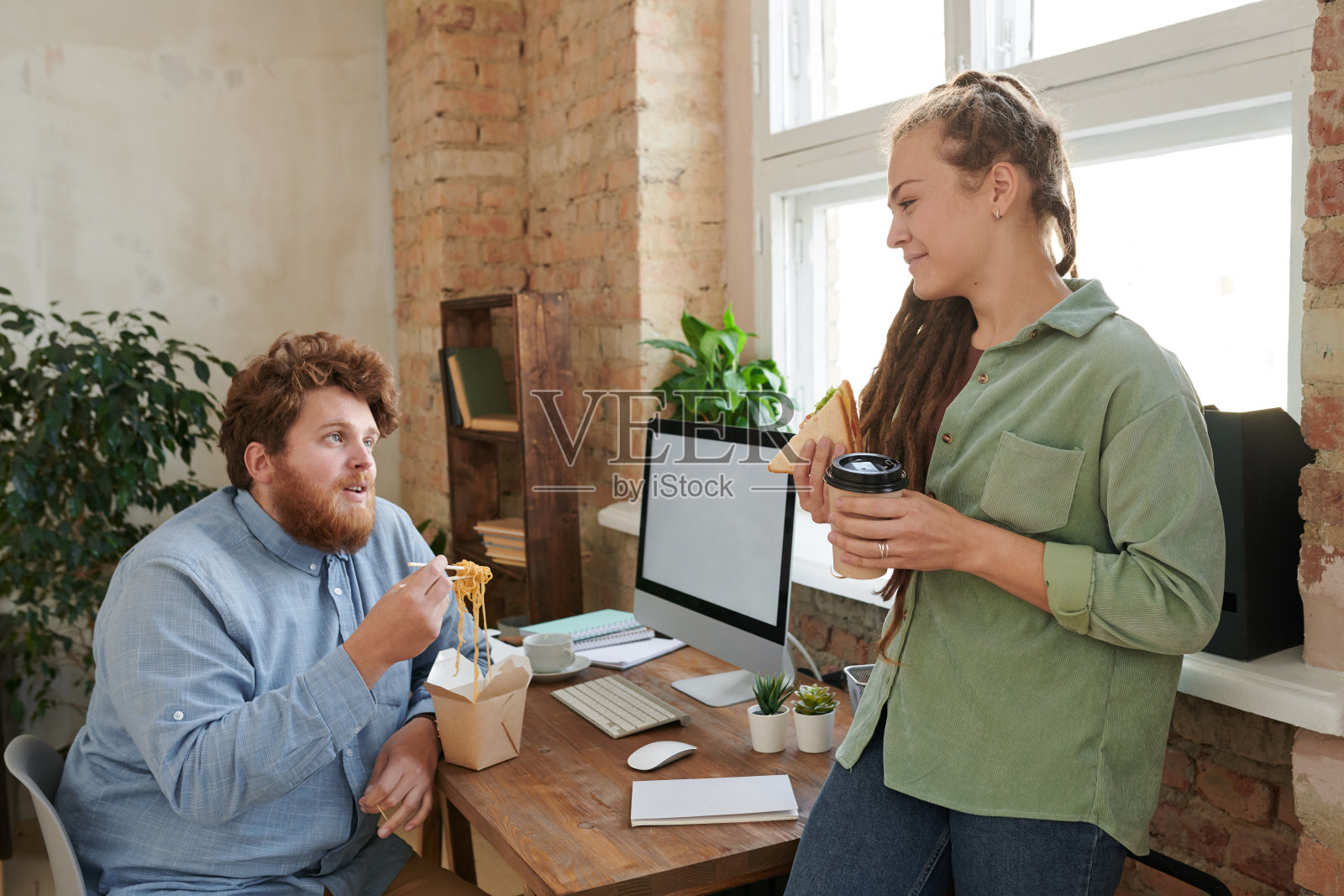 微笑的年轻女子与三明治和咖啡听肥胖的设计师,而他们在工作场所的午餐谈话照片摄影图片