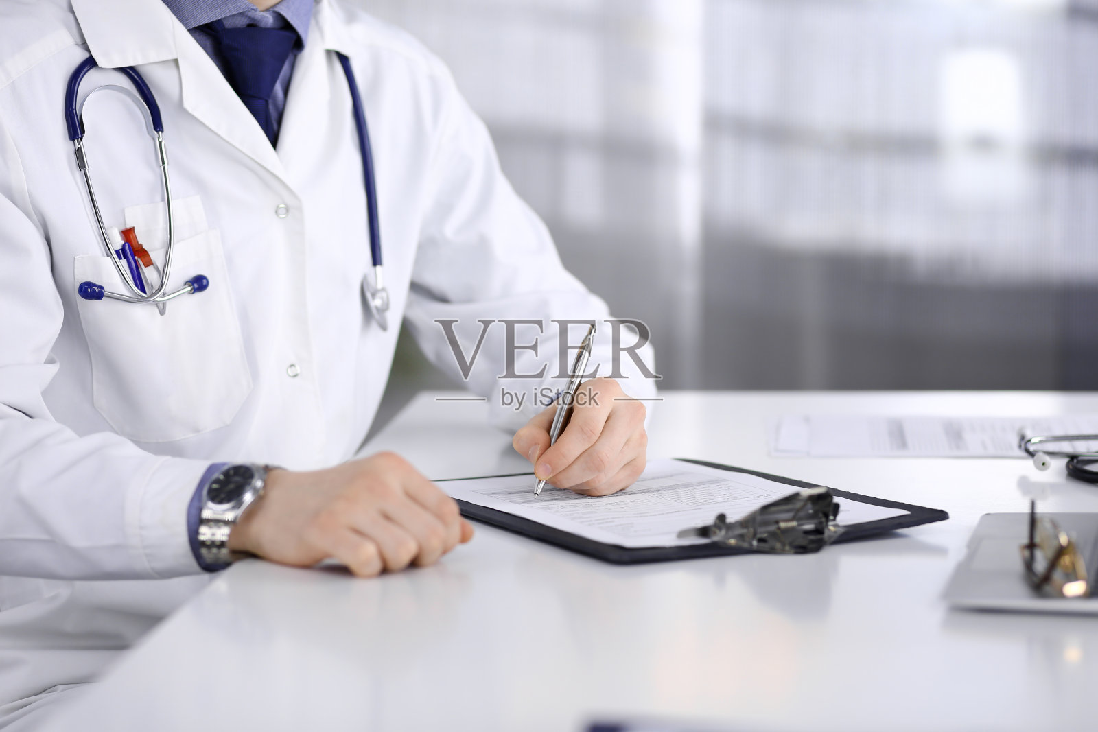 未知男医生在其工作地点的诊所里坐着工作,带着药史记录的剪贴板,特写。年轻医生在工作。完善的医疗服务,医学理念照片摄影图片