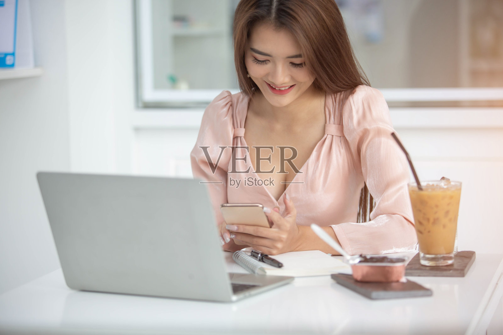 这是一位亚洲商务女性在靠窗的白色桌子上,用电脑、电话和笔记本电脑工作,桌上放着书和咖啡杯。在她的办公室照片摄影图片