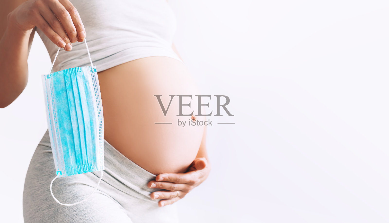 特写孕妇的肚子和医用口罩。大流行期间的保护和预防、孕妇及其婴儿健康的概念。照片摄影图片