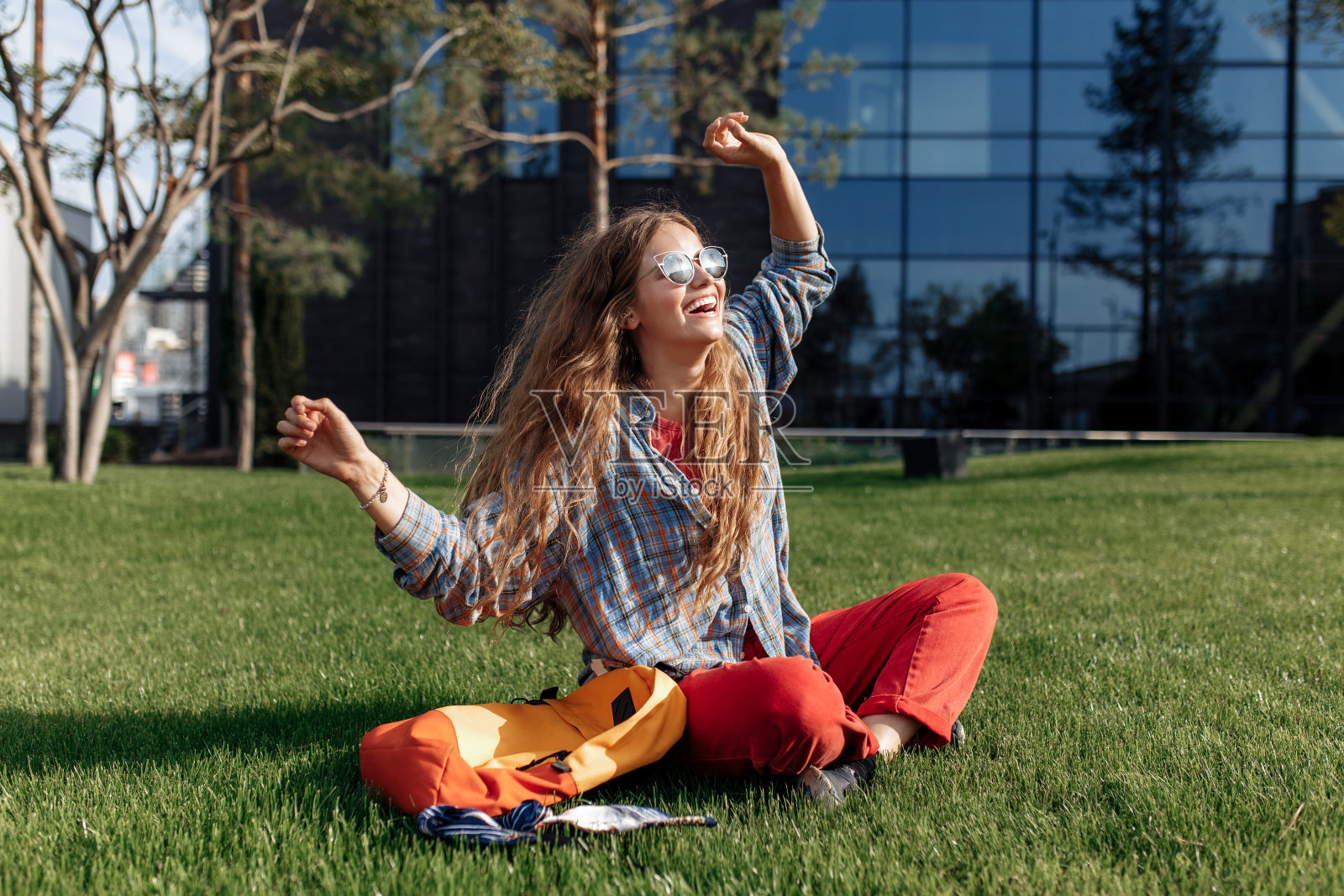 开朗年轻的金发时髦女学生背着背包坐在大学草地上。照片摄影图片