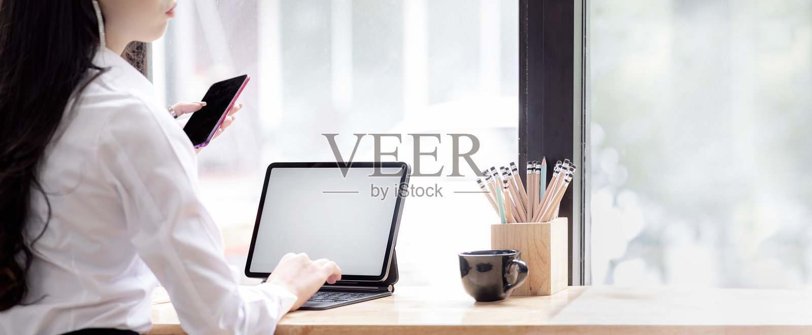 一名身穿白衬衫、手持智能手机、使用数字平板电脑的年轻女子坐在共同工作空间的木制柜台前。照片摄影图片