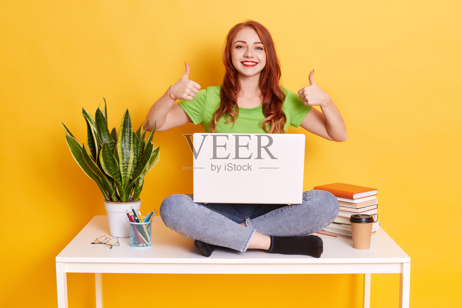 快乐兴奋的女士穿着绿色t恤和牛仔裤,坐在桌子上膝上笔记本电脑,坐在耳朵堆书和花盆,女性显示红色的嘴唇大拇指向上,精神振奋。照片摄影图片