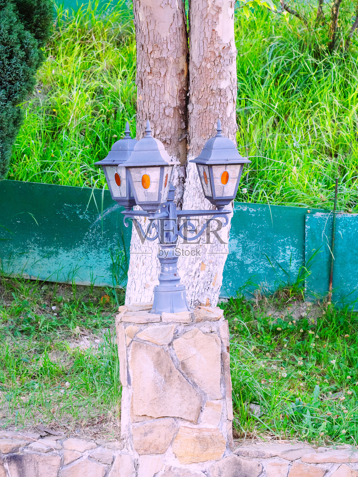 三层装饰街灯在一根短杆和一个石制的支架上,以树干和绿草为背景照片摄影图片