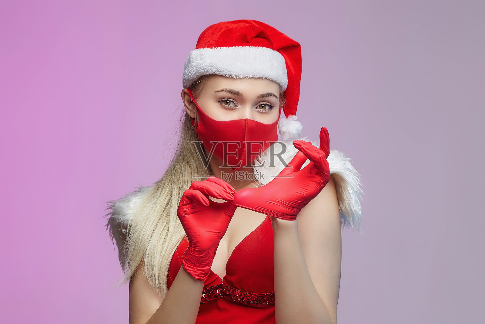 小女孩戴着圣诞老人帽和面具,双手戴着红色手套。圣诞节与COVID-19预防理念照片摄影图片