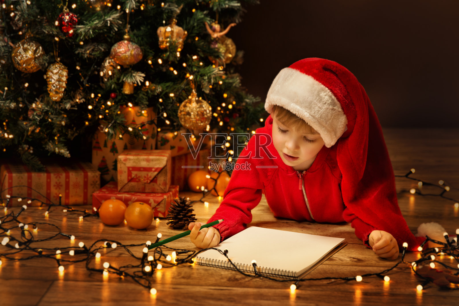 圣诞孩子给圣诞老人写信。孩子在圣诞帽写礼物愿望清单旁边的圣诞树在黑暗的背景照片摄影图片