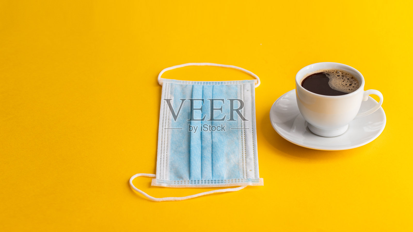 一杯咖啡和一次性口罩上的黄色背景作为新现实的象征。照片摄影图片