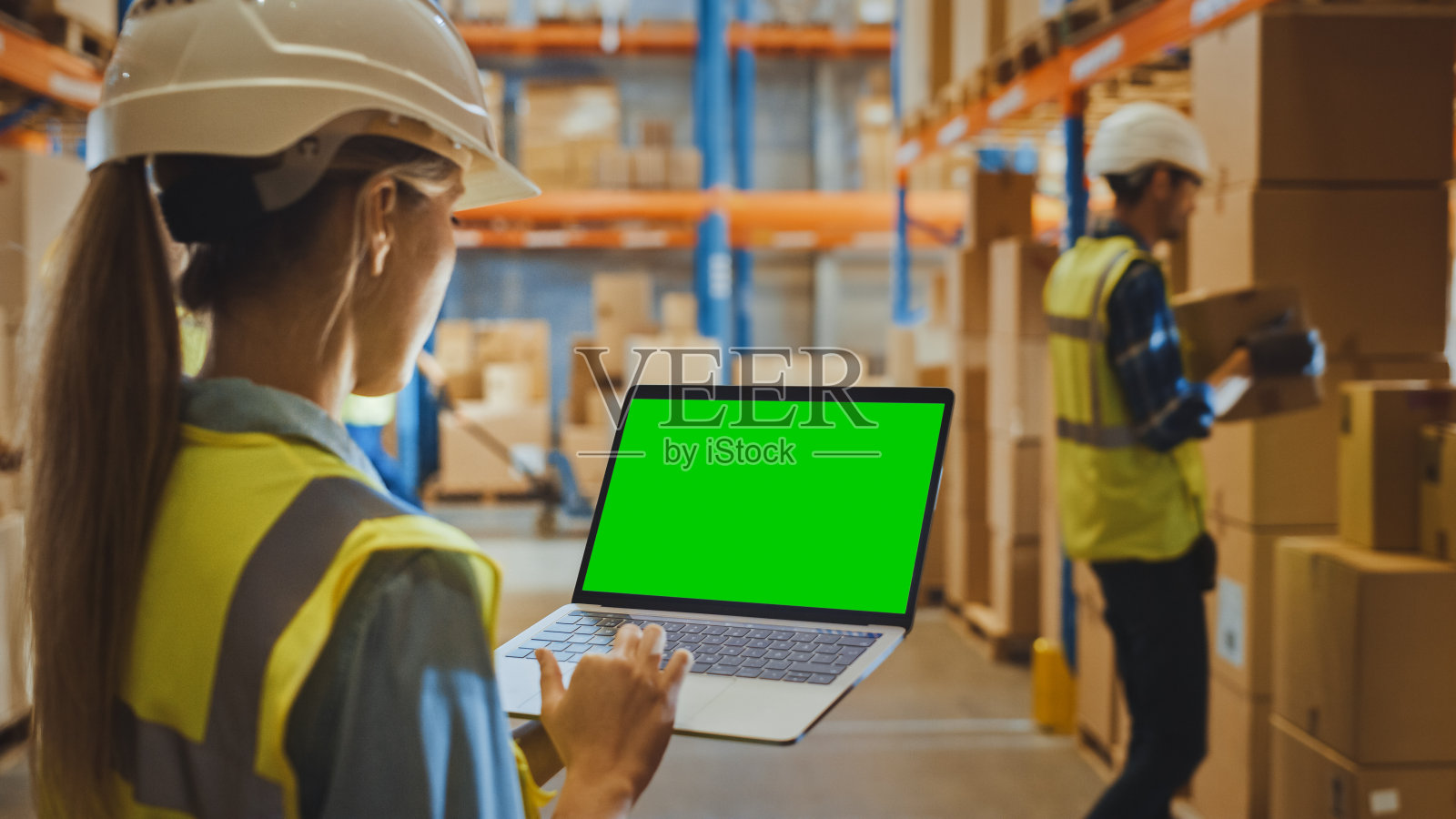 职业女员工戴安全帽,手提电脑绿色色度键屏在景观模式下,在零售仓库堆满了货架和交付货物。侧视图照片摄影图片