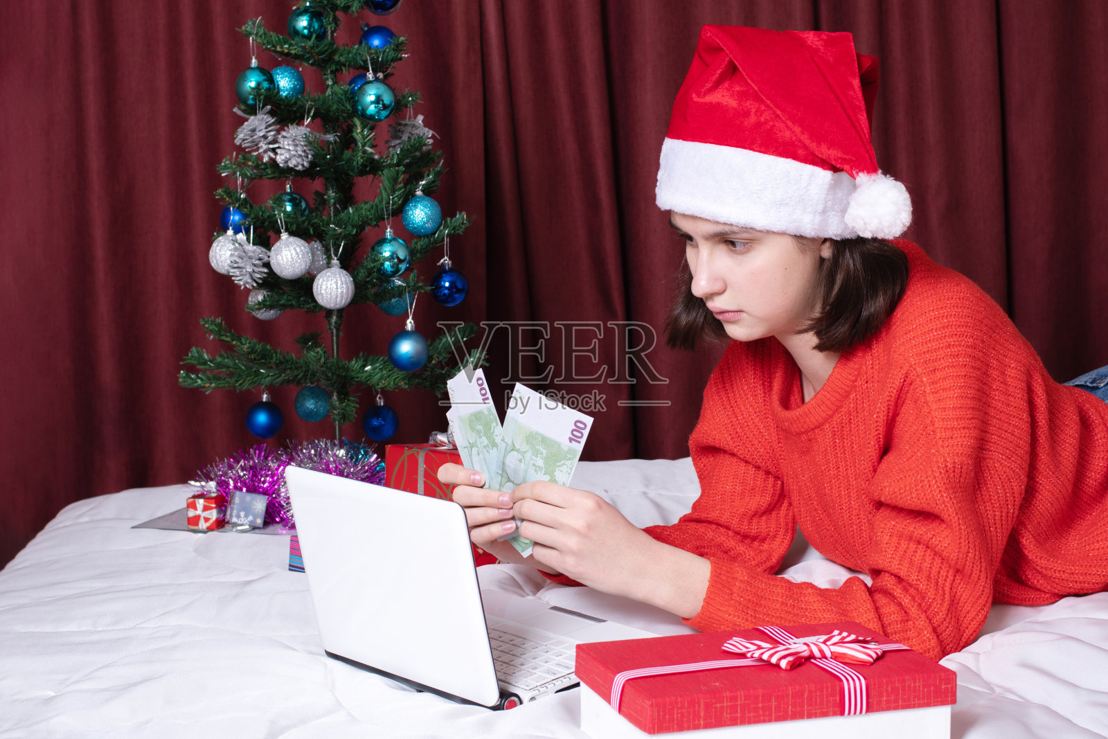 一个白人女孩,戴着一顶红色的圣诞帽,手里拿着一堆现金,看着笔记本电脑照片摄影图片