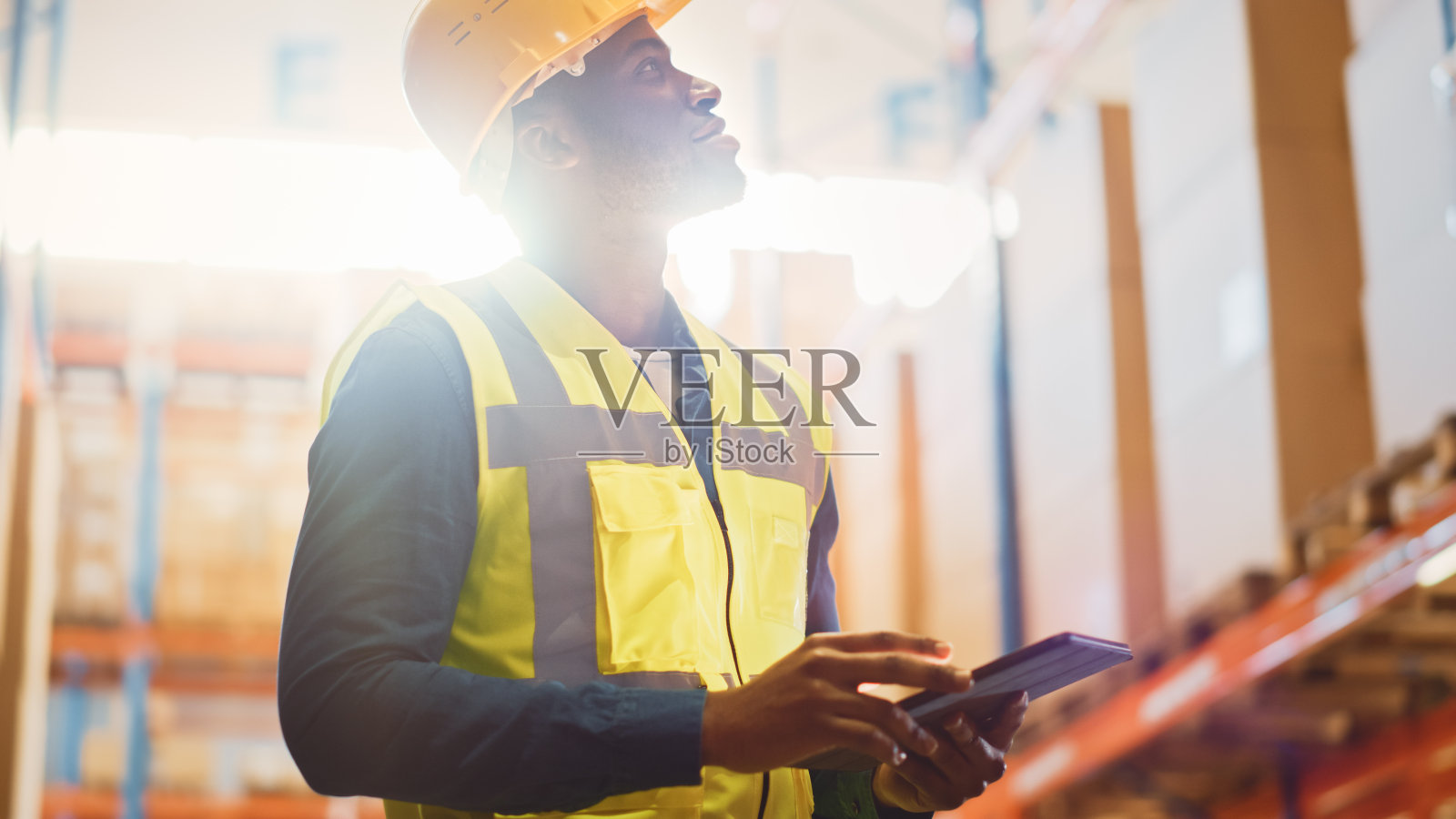 男工人戴安全帽检查产品库存和库存数字平板电脑站在零售仓库满货架的货物。近景低角度拍摄。照片摄影图片