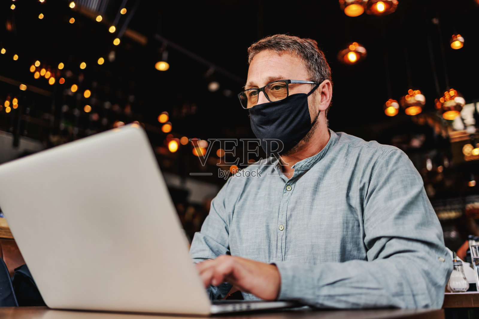 中年严肃的自由职业者戴着眼镜和防护面具坐在咖啡馆里使用笔记本电脑在日冕爆发期间。照片摄影图片