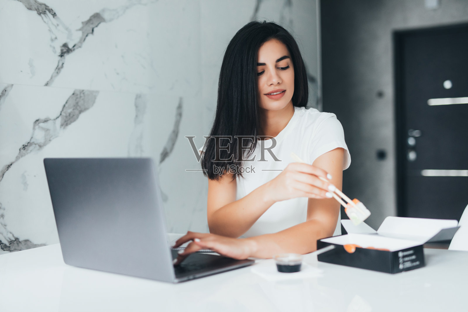 一个年轻的女人坐在她的厨房里吃寿司在家用笔记本电脑工作照片摄影图片