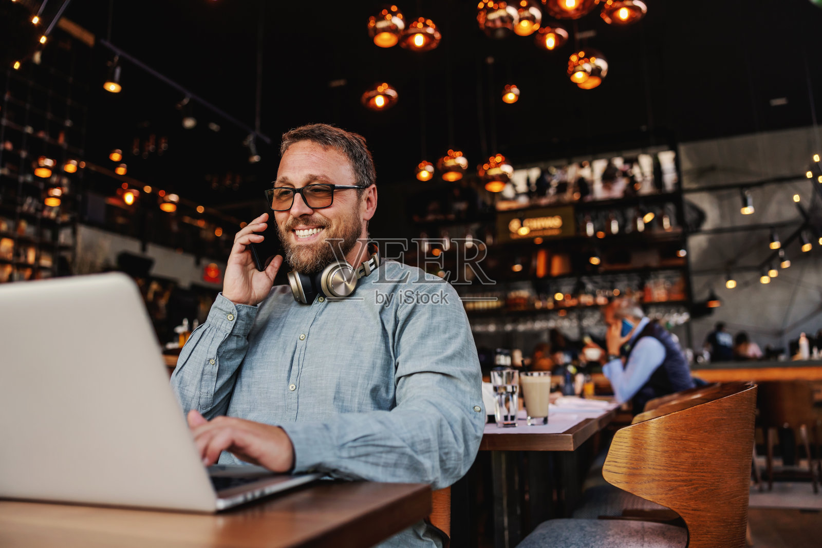 微笑友好的蓄着胡子的中年自由职业者坐在酒吧里,与雇主通电话,使用笔记本电脑工作。照片摄影图片