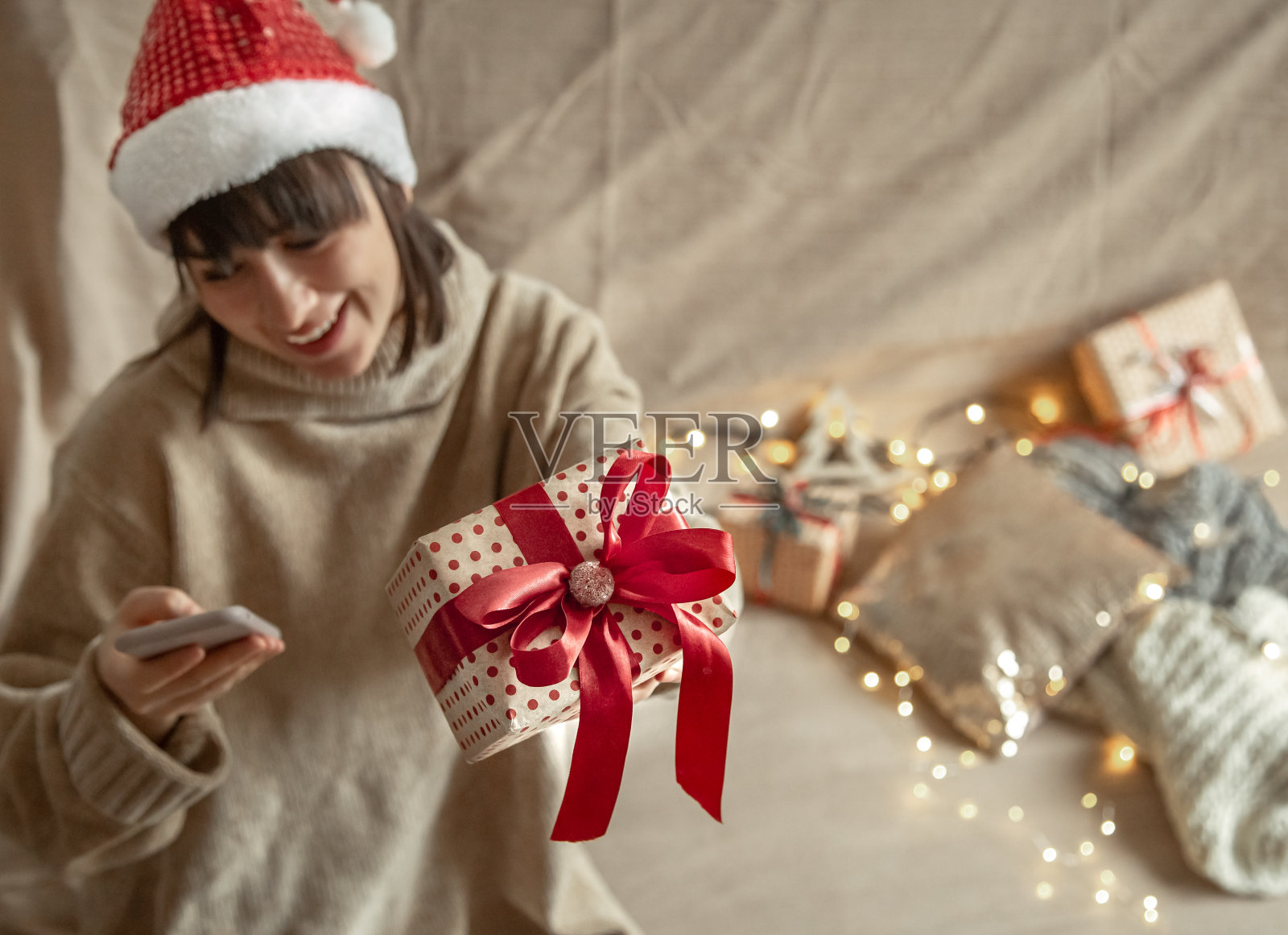 美丽的年轻女子看手机,拿着一个圣诞礼物在模糊的背景。照片摄影图片
