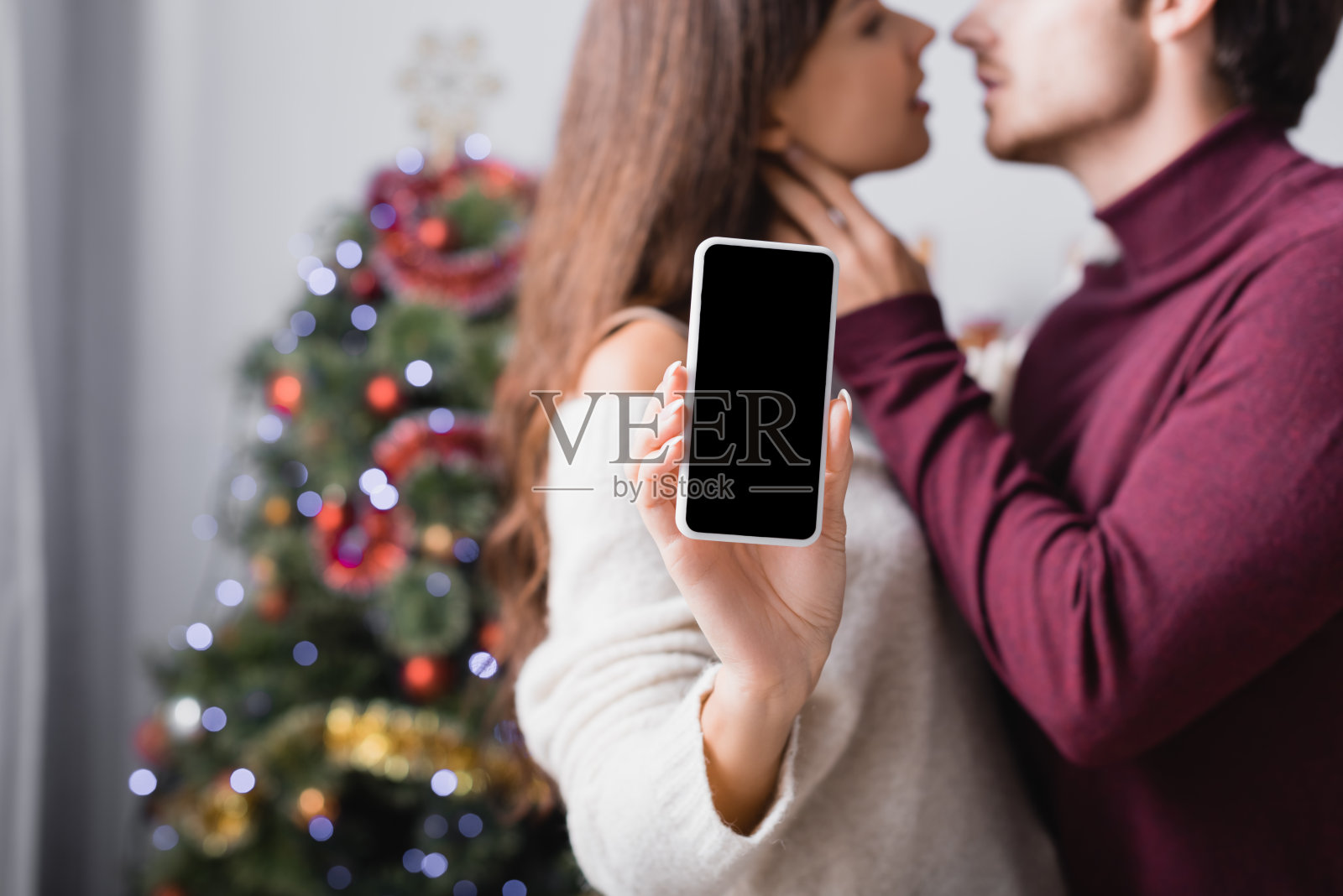 一个女人拿着智能手机,在模糊的背景下,男朋友和圣诞树旁边的空白屏幕照片摄影图片