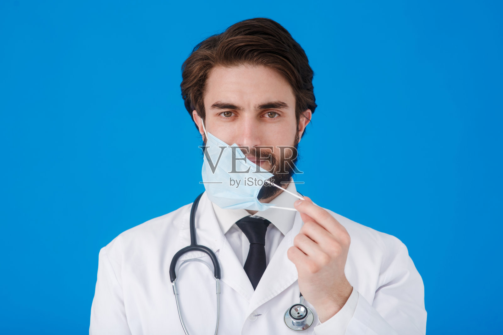医生身穿白大褂,戴着医用口罩,脖子上挂着一个显微镜,背景是蓝色。防范病毒和covid-19的概念照片摄影图片
