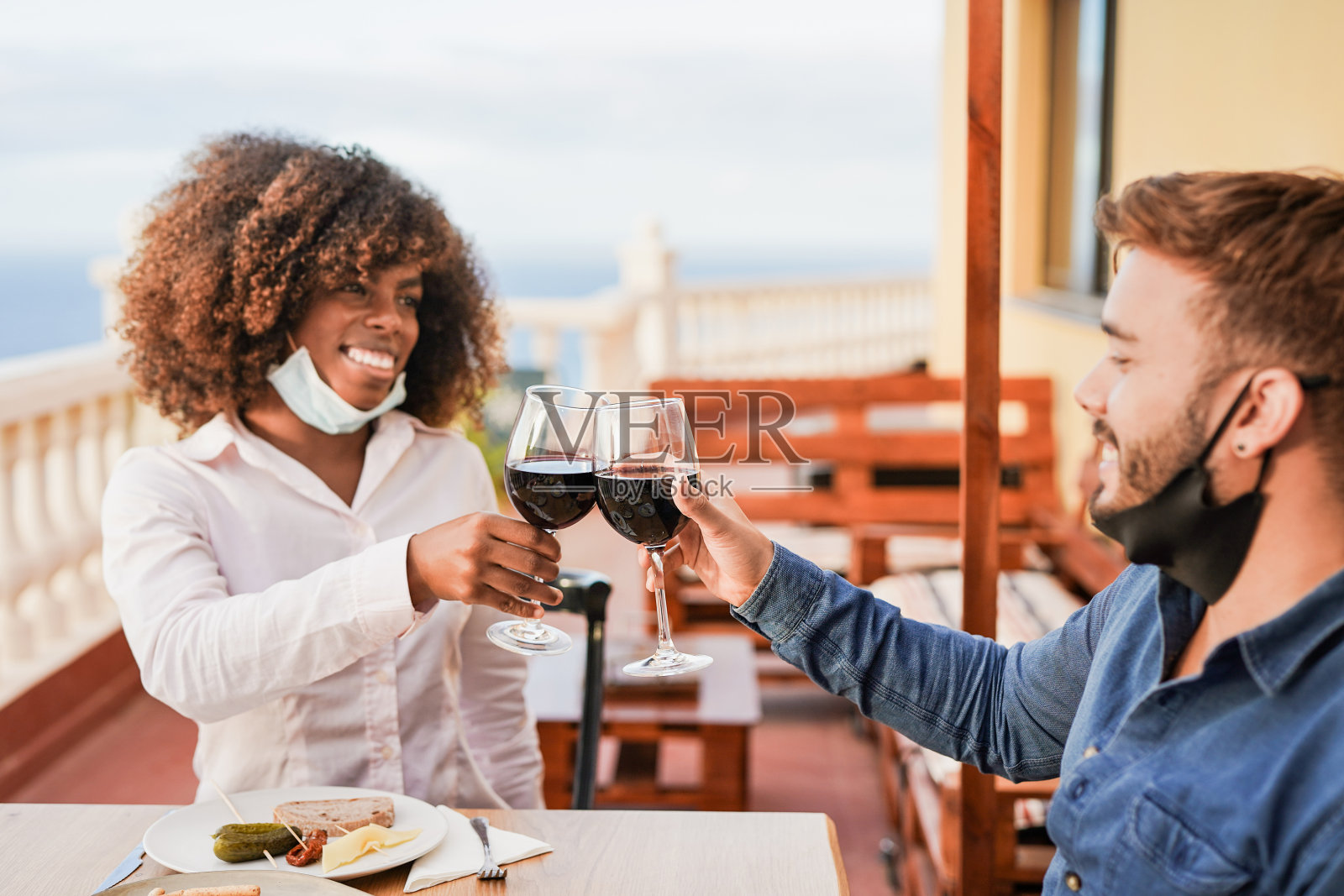 年轻的多种族朋友在家里的露台上喝着红酒欢呼,同时戴着防护面罩照片摄影图片