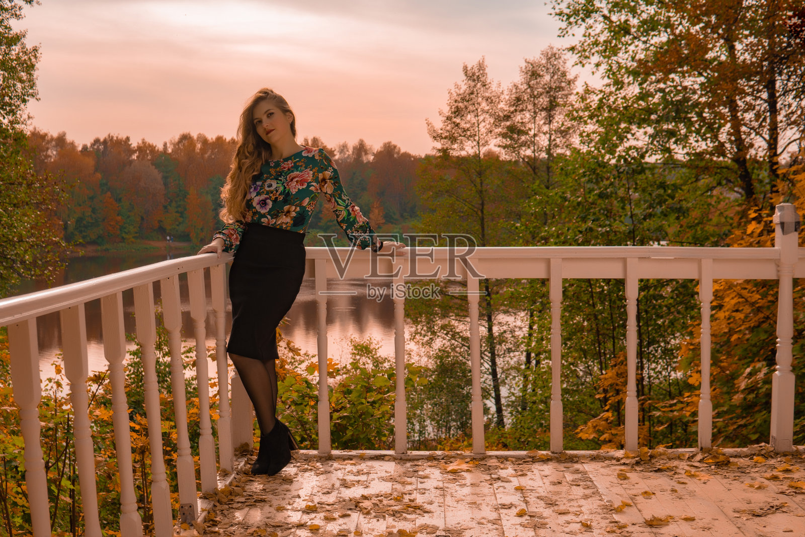 快乐的女子倚着白色的栏杆站在一旁看着镜头,金发碧眼的年轻女子穿着一件彩色的衬衫,在湖水的背景下,天真的秋学院站着一双明亮的蓝眼睛照片摄影图片