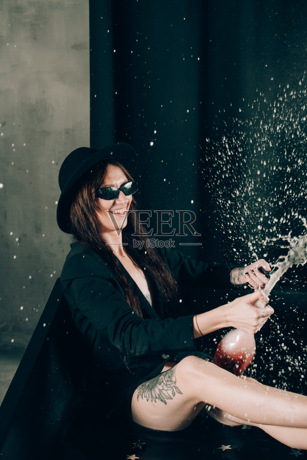 女孩在黑色夹克和帽子摆姿势在黑色浴缸。一瓶葡萄酒。手里拿着枪照片摄影图片