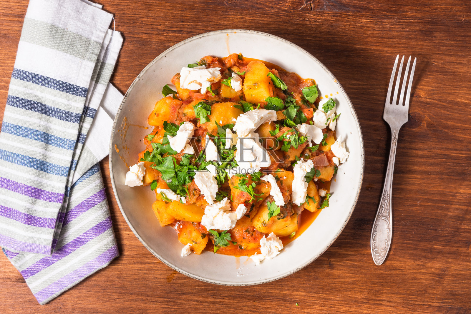 希腊蔬菜炖土豆和蔬菜在番茄酱和软羊乳酪块在一个盘子里放在木桌上,旁边有一把叉子和餐巾,俯视图照片摄影图片