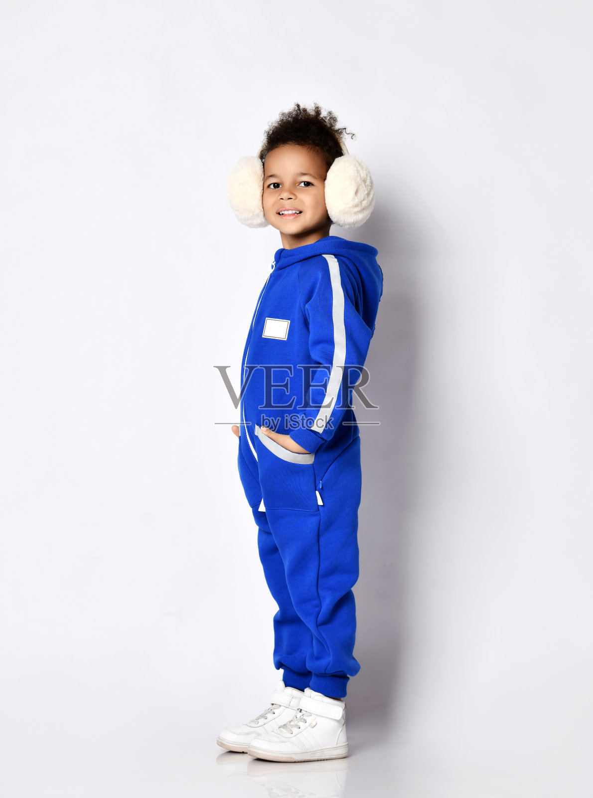 快乐微笑的黑皮肤的小女孩在温暖的蓝色时尚连衣裤和白色运动鞋站在相机的一边照片摄影图片