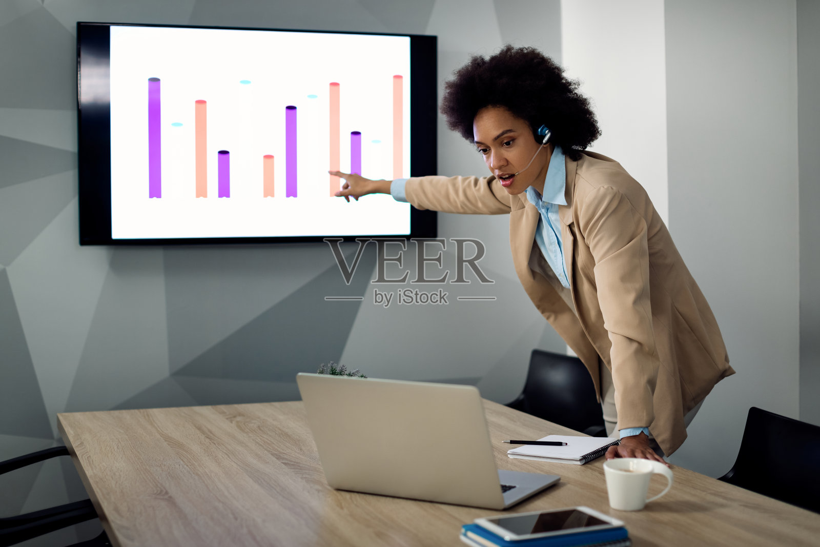 非裔美国女商人正在用笔记本电脑进行电话会议,并指着办公室投影屏幕上的图表。照片摄影图片