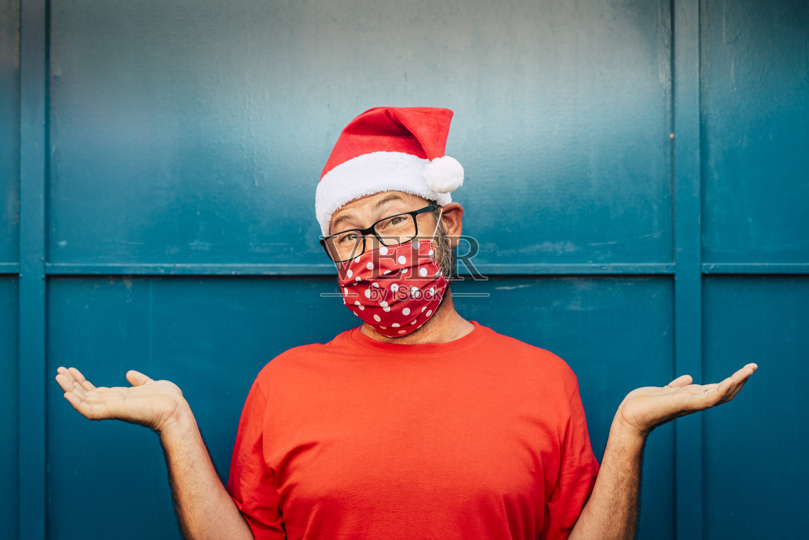 在冠状病毒新冠肺炎紧急情况下,戴着红色圣诞帽和口罩的快乐而孤独的成年男子肖像——封锁期间的圣诞和假日概念照片摄影图片