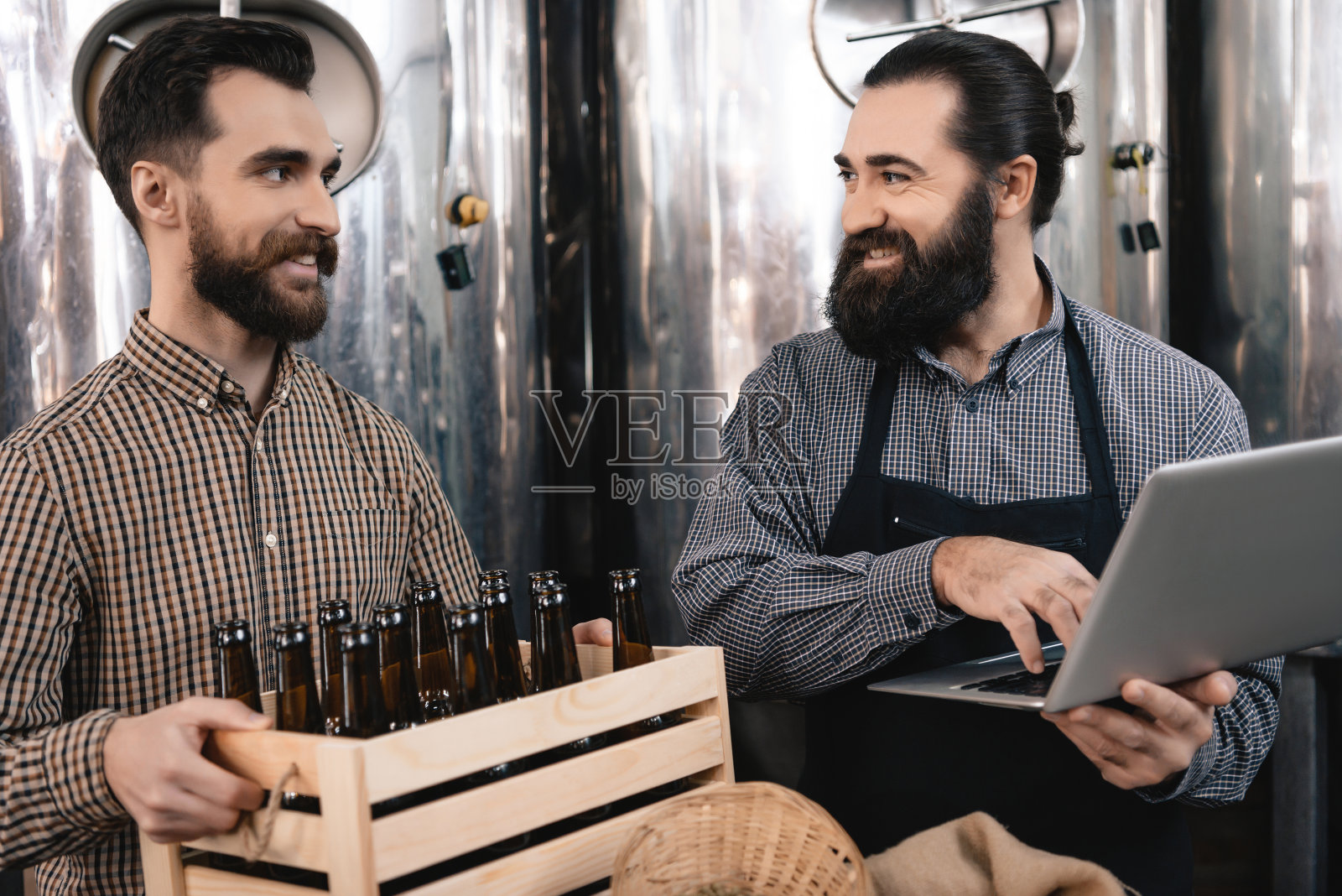 穿着围裙、长胡子的酿酒人看着快乐的男人,手里拿着一箱空瓶子。照片摄影图片